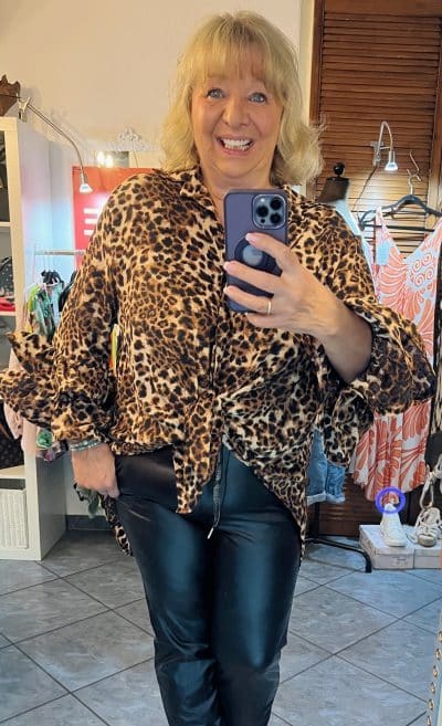 Eine Person macht ein Spiegel-Selfie in einem mit verschiedenen Gegenständen dekorierten Raum. Sie trägt eine Bluse mit Leopardenmuster und die zauberhafte LeatherStyle Hose von GlamGirl. Die Person lächelt, während sie ein Smartphone hält und ihr Spiegelbild einfängt.