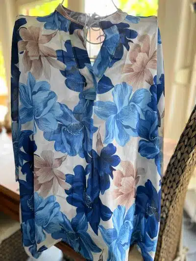 Eine Bloom Chic Bluse mit Blumenmuster hängt über einem Stuhl. Große Blumen in Blau- und Brauntönen zieren den weißen Hintergrund und bilden ein auffälliges Design. Sie verfügt über einen Kragen und eine teilweise geknöpfte Vorderseite und verbindet mühelos Stil und Komfort.