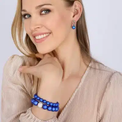Eine Frau mit langen blonden Haaren und einer beigefarbenen Bluse lächelt in die Kamera. Sie trägt blaue Ohrringe und ein passendes Magic Beads CC-Armband (Kopie) mit einer silbernen Perle. Sie stützt ihr Kinn auf ihre rechte Hand und zeigt das Armband.