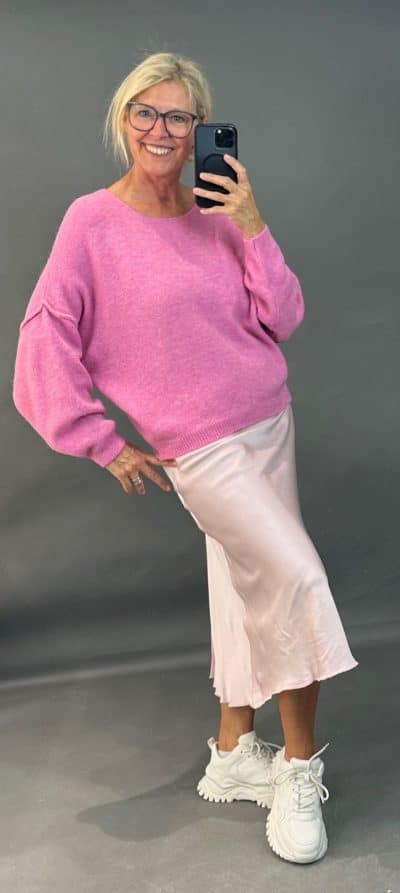 Eine Frau posiert für ein Selfie und lächelt in die Kamera. Sie trägt eine Brille, einen rosa Urban Cozy Pulli und Pullover, einen hellrosa Rock und weiße Turnschuhe. Sie hält ihr Telefon in der rechten Hand und hat ihre linke Hand auf der Hüfte. Der gemütliche Hintergrund ist ein schlichtes Dunkelgrau.