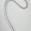Ein Bild zeigt die magische Wechselkette, eine lange doppelreihige Halskette, die mit weißen Perlen verziert ist und einen runden Silberverschluss mit kleinen eingelegten Steinen aufweist. Die Kette ist elegant vor einem schlichten weißen Hintergrund angeordnet, was ihre raffinierte Schlichtheit betont.