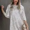 Eine Frau mit langem, welligem Haar steht vor einer dunklen Wand. Sie trägt ein silbernes Paillettenkleid. In einer Hand hält sie eine beige Clutch, trägt roten Lippenstift und blickt direkt in die Kamera.