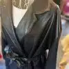 Eine Nahaufnahme zeigt den Black Fashion Trench, einen modernen Kunstledermantel, der elegant über eine Drahtpuppe drapiert ist. Er verfügt über einen stilvollen Reverskragen und wird durch einen passenden Gürtel um die Taille betont. Ein weißes Kleidungsstück darunter sorgt für einen auffälligen Kontrast zum Gesamtlook.