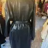Der Black Fashion Trench wird elegant auf einer Schaufensterpuppe im Geschäft präsentiert, sein glänzendes Lederfinish fällt ins Auge. Dieser schicke Trenchcoat hat einen Gürtel in der Taille, im Hintergrund sind auf Ständern eine Auswahl an modischen Kleidungsstücken angeordnet.