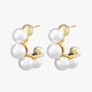 Die Perlencreolen-Ohrringe bestehen aus runden Ohrsteckern aus Gold, die mit Gruppen weißer Perlen verziert sind, die anmutig in einer Kurve angeordnet sind und einen stilvollen und eleganten Kontrast zu einem schlichten, hellen Hintergrund bilden