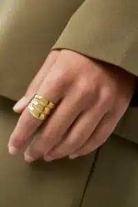 Eine Hand mit einem goldenen Dreibandring ruht auf einer Oberfläche. Die Person trägt ein grünes langärmeliges Kleidungsstück. Der Hintergrund ist verschwommen und passt farblich zur Kleidung