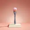 Der Beauty Queen-Make-up-Pinsel mit rosa Borstenkopf und einem mit bunten Perlen verzierten Griff steht elegant auf einem zweistufigen beigen Sockel vor einem pfirsichfarbenen Hintergrund.