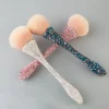 Drei Beauty Queen-Make-up-Pinsel mit flauschigen Borsten und glitzernden, mit Strasssteinen besetzten Griffen in Silber, Pink und Blaugrün sind elegant kreuz und quer auf einem hellgrauen Hintergrund angeordnet, um ihre wahre Schönheit zur Geltung zu bringen.