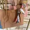 Hol dir den ultimativen Style-Kick mit dieser bezaubernden, beigefarbenen Handtasche! Die tan-farbenen Griffe harmonieren perfekt mit dem lebendigen Pink des gemusterten Tuchs – ein echtes Fashion-Statement! Im Hintergrund blitzt eine schicke pinke Tasche hervor und sorgt für noch mehr Glamour. Beide Taschen hängen an einem eleganten Metallständer vor einer stylischen Steinstrahlwand. Zeit, deinem Outfit das gewisse Etwas zu verleihen!.