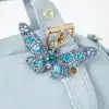 Lust auf ein echtes Statement-Piece? Die Butterfly Handbag in zartem Blau wartet auf dich! Goldene Hardware verleiht ihr einen edlen Twist, während die funkelnde Schmetterlings-Brosche mit blauen und türkisen Strassteinen direkt am Griff alle Blicke auf sich zieht. Das perfekte Accessoire, wenn du deinen Style aufs nächste Level heben willst – bist du bereit zu strahlen?.