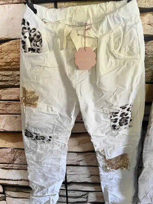Mach dich bereit für deinen neuen Lieblings-Look: Die zauberhafte Patch-Hose ist wie für dich gemacht! Die edle weiße Crinkle-Optik trifft auf funkelnde goldene Pailletten und coole Leo-Print-Patches - ein echter Eyecatcher, der garantiert alle Blicke auf dich zieht. Am lässigen Backstein-Hintergrund wirkt dein Style urban, aufregend und absolut einzigartig. Wage den Trend und bringe Glamour in deinen Alltag!.