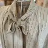 Erlebe mit der Line Style Tunika pure Eleganz - eine beige Nadelstreifen-Bluse, die mit feinen Streifen und einem charmanten Schleifenkragen alle Blicke auf sich zieht. Der raffinierte Schnitt bringt deine Silhouette auf moderne Weise zur Geltung und verleiht dir einen individuell-stylischen Look. Auf einem weißen Draht-Kleiderständer präsentiert, inspiriert die Tunika dich, deinen persönlichen Stil neu zu definieren - zeitlos schön, vielseitig kombinierbar und immer ein Fashion-Statement. Starte jetzt durch und mache dieses Trend-Piece zum Mittelpunkt deines Outfits!.