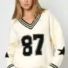 Entdecke deinen Style: Im cremefarbenen Varsity Star Pulli mit coolen Streifen, Nummer 87 und Stern-Designs setzt du Trends.