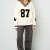 Zeig deinen Style mit dem cremefarbenen Varsity Star Pulli, grauen Jeans und coolen Sneakern vor cleanem, weißem Hintergrund.