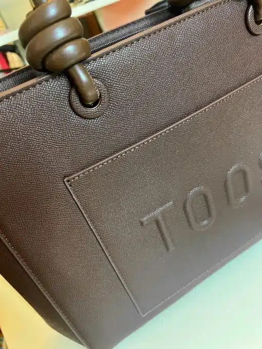 Erlebe das moderne Design der "Toos" Handtasche: edle braune Textur, runde Griffe und stilvoll geprägtes Logo.