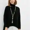 Setze ein Statement: Du trägst einen schwarzen Rollkragenpullover, weiße Hosen und eine lange, trendige Kette mit Quaste.