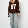 Style den angesagten Varsity Star Pulli in Braun mit "87" und deinen hellblauen Jeans für einen freshen Look vor weißer Wand.