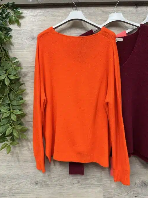 Entdecke deinen neuen Lieblingspullover: Ein leuchtender oranger Sweater neben einem burgundroten Akzent an einer Holz-Wand.