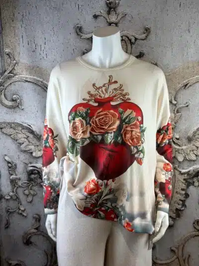 Du trägst einen angesagten Creme-Pullover mit auffälligem Sacred-Heart- und Rosenmotiv auf Front und Ärmeln.