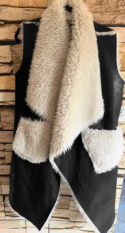 Erlebe deinen Style neu: Die schwarze Urban Chic Wendeweste mit weißem Webpelzkragen setzt dich vor Steinwand perfekt in Szene.