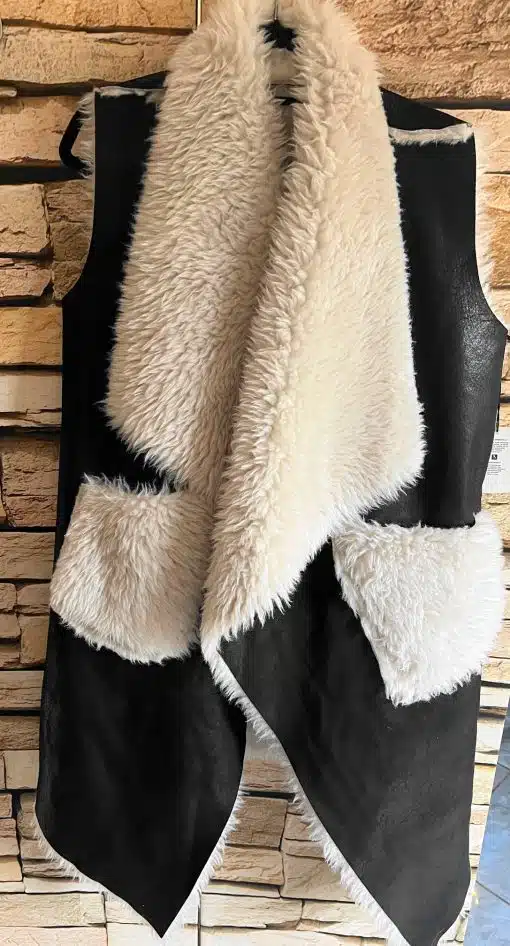 Erlebe deinen Style neu: Die schwarze Urban Chic Wendeweste mit weißem Webpelzkragen setzt dich vor Steinwand perfekt in Szene.