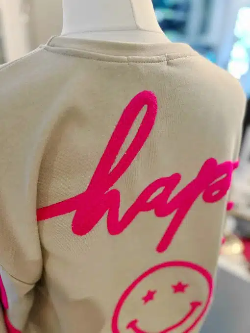 Hol dir das moderne Be Happy Sweatshirt in Beige mit knalligem pinken "hap"-Schriftzug und coolem Smiley mit Sternaugen auf dem Rücken!.