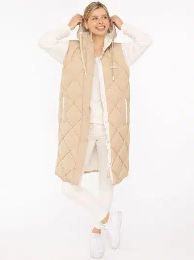 Du trägst eine lange beige Steppweste über weißem Outfit, lächelst und hältst cool die Kapuze - bereit für deinen modernen Look.