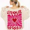 Du trägst einen cremefarbenen Pullover mit auffälligem pink-rotem Sunburst und stylischem "LE SOLEIL"-Schriftzug auf dem Rücken.