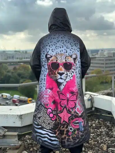 Du trägst einen stylischen Kapuzenmantel mit buntem Geparden- und Pink-Design, stehst cool auf einem Rooftop und blickst hinaus.