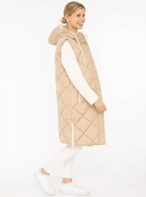 Du trägst eine lange, beige Steppweste über weißem Longsleeve, weiße Hose und Sneaker - modern, lässig, stilsicher.