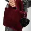 Du hältst eine bordeauxrote Faux-Fur-Handtasche mit schwarzem Plüsch-Liebesanhänger zu deinem coolen Lederlook.
