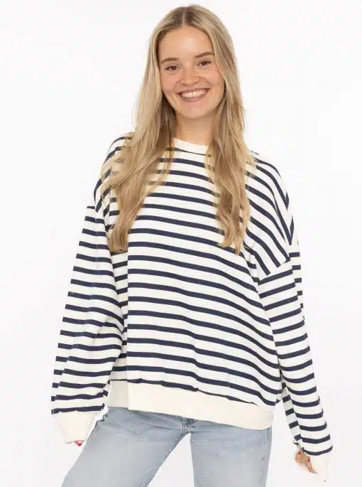 Zeige dein strahlendes Lächeln in einem modernen weißen Pullover mit Navy-Streifen und hellblauen Jeans vor weißem Hintergrund.