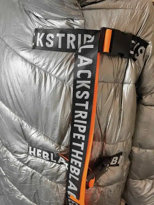 Erlebe das moderne Statement: Silberne Pufferjacke mit BLACKSTRIPE-Trägern und auffälliger, orangefarbener Akzent-Schnalle.