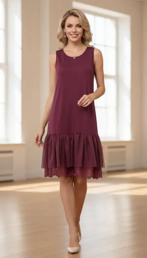 Du trägst ein ärmelloses, bordeauxrotes Layering-Kleid mit Rüschen, stilvoll inszeniert vor großen Fenstern im hellen Raum.