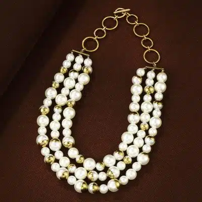 Entdecke dein neues Must-have: Dreireihige Perlen-Goldkette - ein echtes Statement auf braunem, stilvollem Hintergrund.