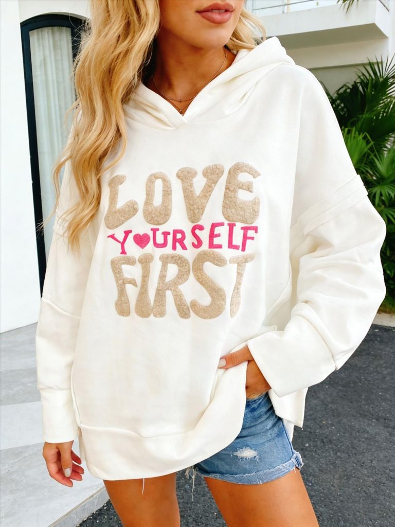 Eine Frau modelliert den weißen Yourself-Kapuzenpulli mit der Aufschrift LOVE YOURSELF FIRST" (LIEBE DICH SELBST ZUERST) in Hellbraun/Pink, der Komfort, Stil und Selbstvertrauen demonstriert.