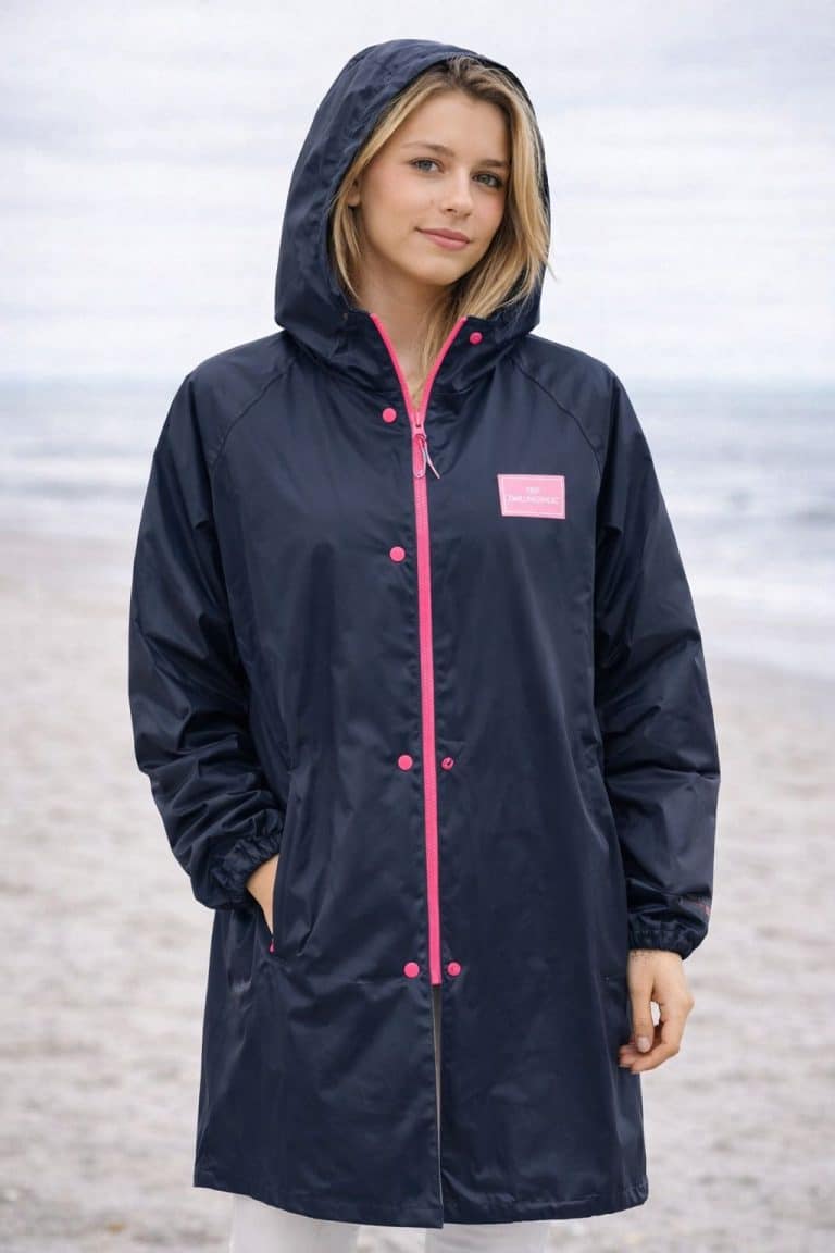 Das Model trägt einen langen marineblauen Rain Uni Mantel von Zwillingsherz mit Kapuze, pinkfarbenen Akzenten, Brustaufnäher und Druckknöpfen - ein stylischer Regenschutz.