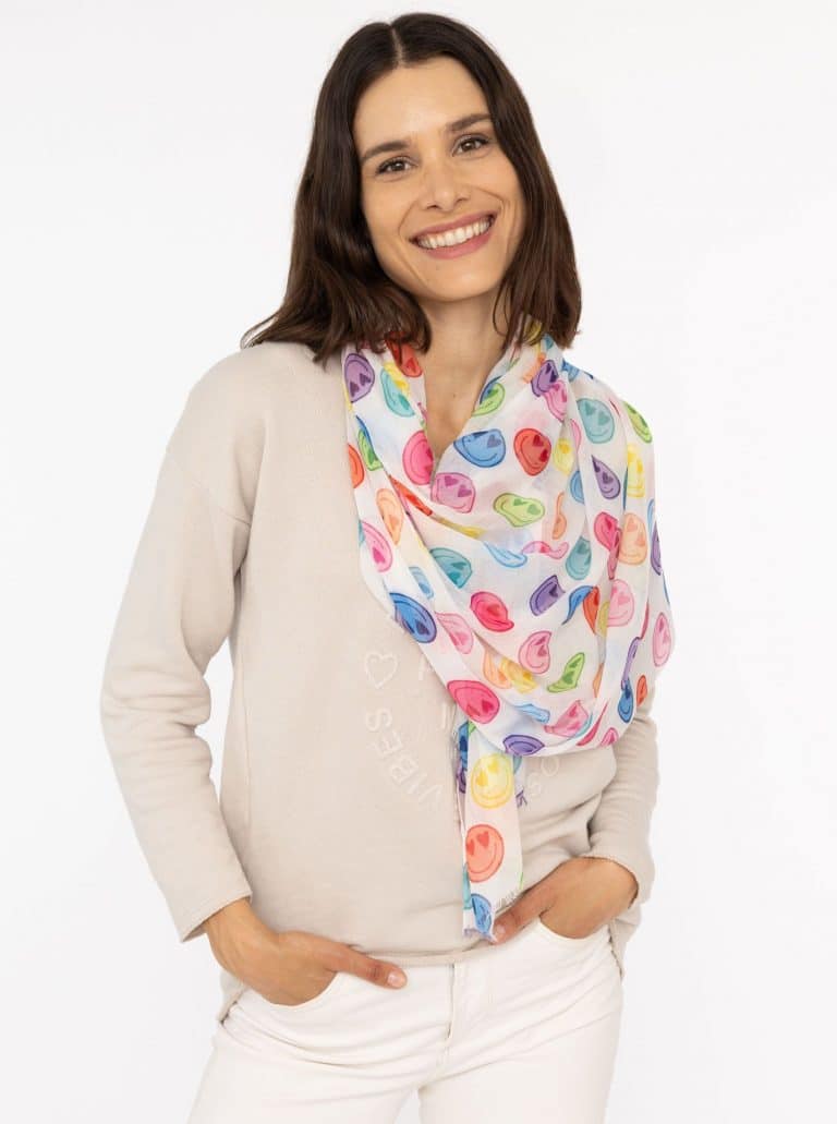 Eine Frau modelliert einen beigen Pullover und eine weiße Hose mit einem leuchtenden Smiley-Schal, der jedem Outfit einen lustigen Touch verleiht; der Hintergrund ist schlicht weiß.