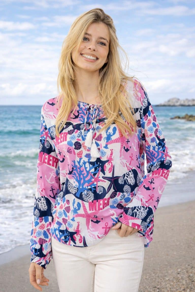 Frau am Strand in farbenfroher Maritime Mood Tunikabluse und weißer Hose - lebendiger, stilvoller Komfort, perfekt für Tage am Meer.