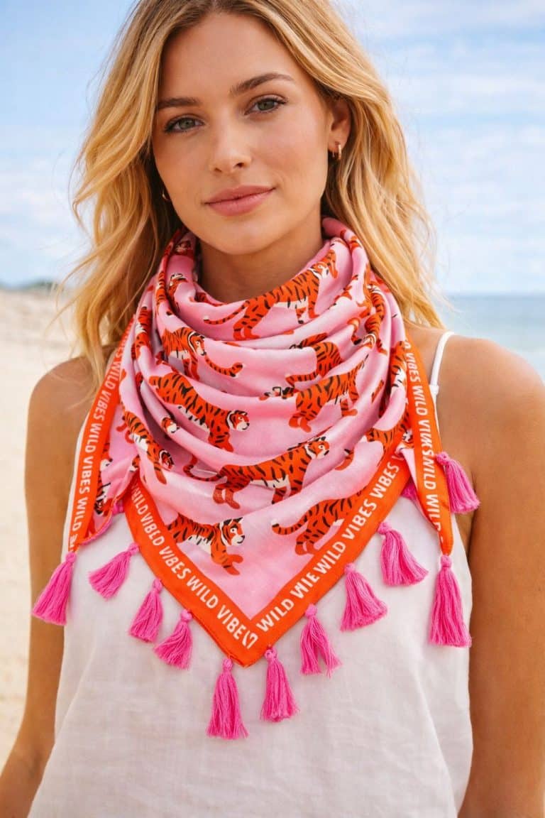 Blonde Frau am Strand trägt Zwillingsherz Beach Vibes Schal mit orangem Tiger-Print, roter Borte und rosa Quasten - mutiger Stil und Sommerflair.