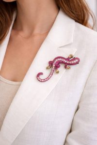 Das Model trägt einen weißen Blazer und ein beiges Oberteil mit der Geko Brosche, einer auffälligen rosa Schmuckbrosche, die jedem Outfit Eleganz und Luxus verleiht.