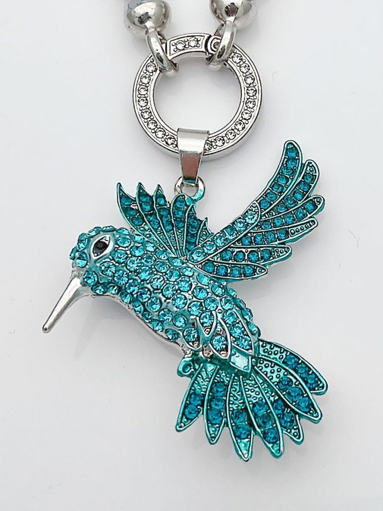 Kolibri Wechsel Anhänger: A close-up shows a hummingbird pendant with blue rhinestones on a silver chain, shimmering brightly.