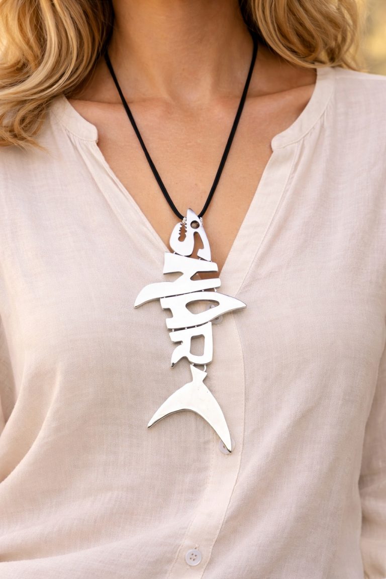 Close-up of the Shark Kette, a silver shark-shaped pendant on a black cord. The design incorporates the word 'SHARK' into the shark's bone structure.