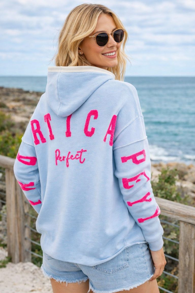 Frau mit Zwillingsherz-Sonnenbrille und blauem Stickerei-Hoodie mit rosafarbenen Details, "Perfekt" im Rücken - stylisch, bequem, ozeantauglich.