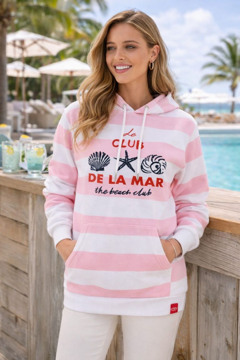 Lächelnde Frau am Pool trägt einen rosa-weiß gestreiften Beach Club Hoodie mit Taschen, Muschelgrafik und "Le Club De La Mar"-Schriftzug - die perfekte, gemütliche Strandkleidung.