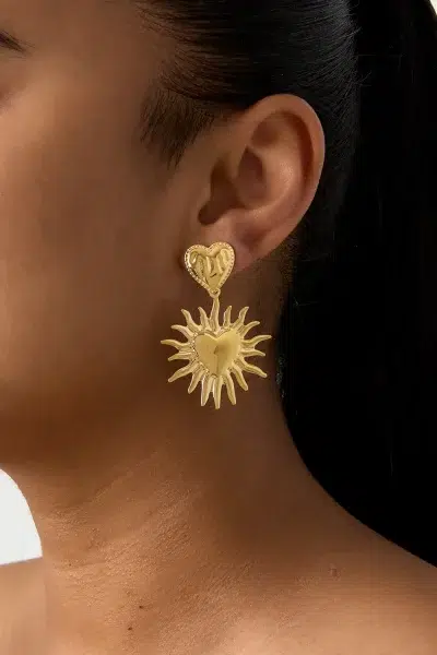 Nahaufnahme eines Models, das Sonnenherz-Ohrstecker trägt: goldfarbene Ohrringe in Herz- und Sonnenstrahlform für einen auffälligen Look und elegante Vielseitigkeit.