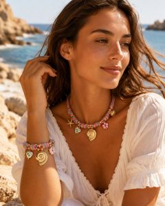 Eine Frau am Meer mit einem „Costa Bella“-Armband mit bunten Perlen, einer goldenen Muschel, Seesternen und Blumenanhängern – das perfekte Sommeraccessoire.
