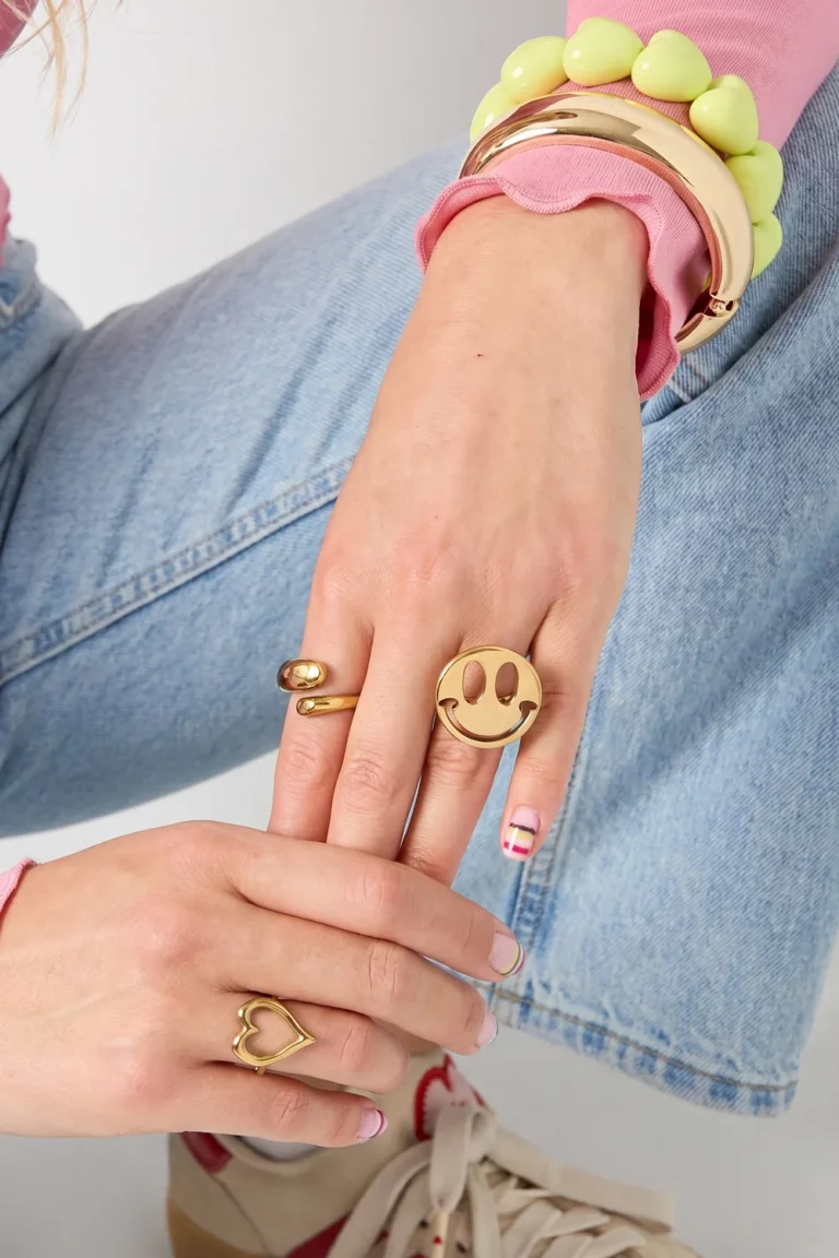 Ein Model in hellblauen Jeans und einem rosa Oberteil präsentiert einen goldenen Smiley-Statement-Ring und ein klobiges Perlenarmband – damit wird jeder Look verspielter.