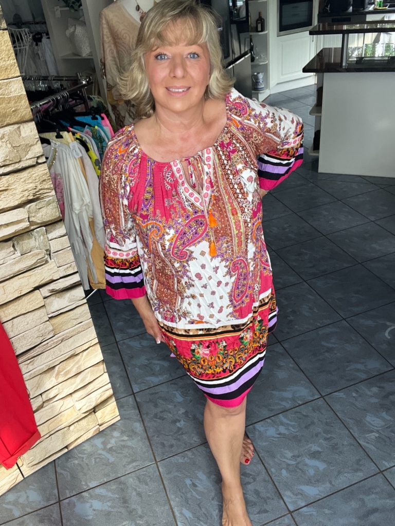 Eine Frau präsentiert das „Fiesta Vibes Tunika-Kleid“ mit lebhaften Mustern in Pink, Orange und Weiß – perfekt für einen lässigen Sommerlook.
