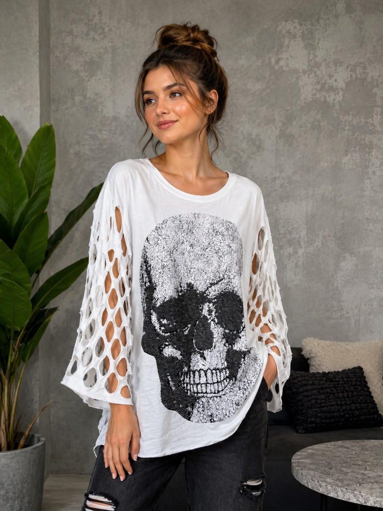 Trendiges weißes Damen-Top mit stilvollen ovalen Ausschnitten an den Ärmeln und auffälligem schwarzem Totenkopf-Print, kombiniert mit einer dunklen Jeans mit Rissen für einen ausgefallenen Look.